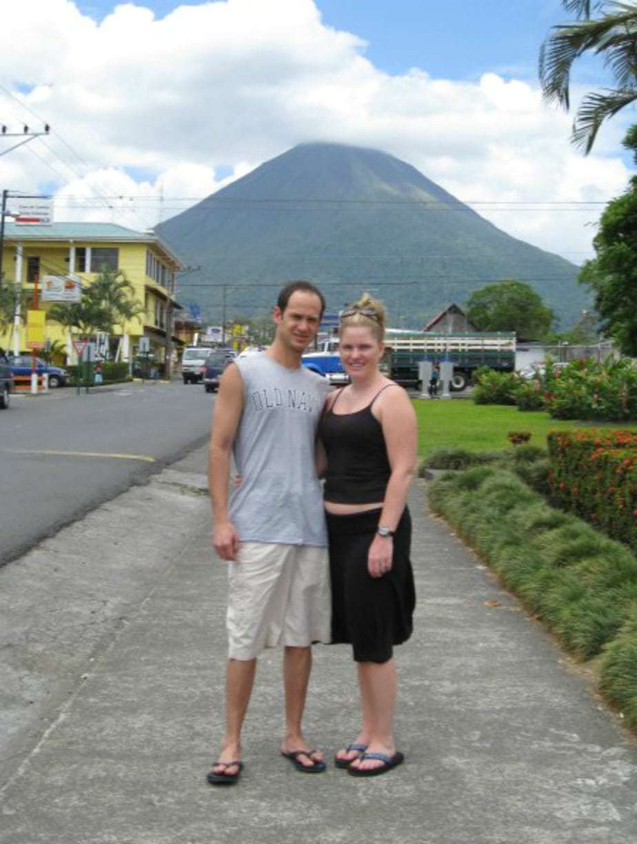 Couple standing together on a sidewalk with the arenal volcano in the background.