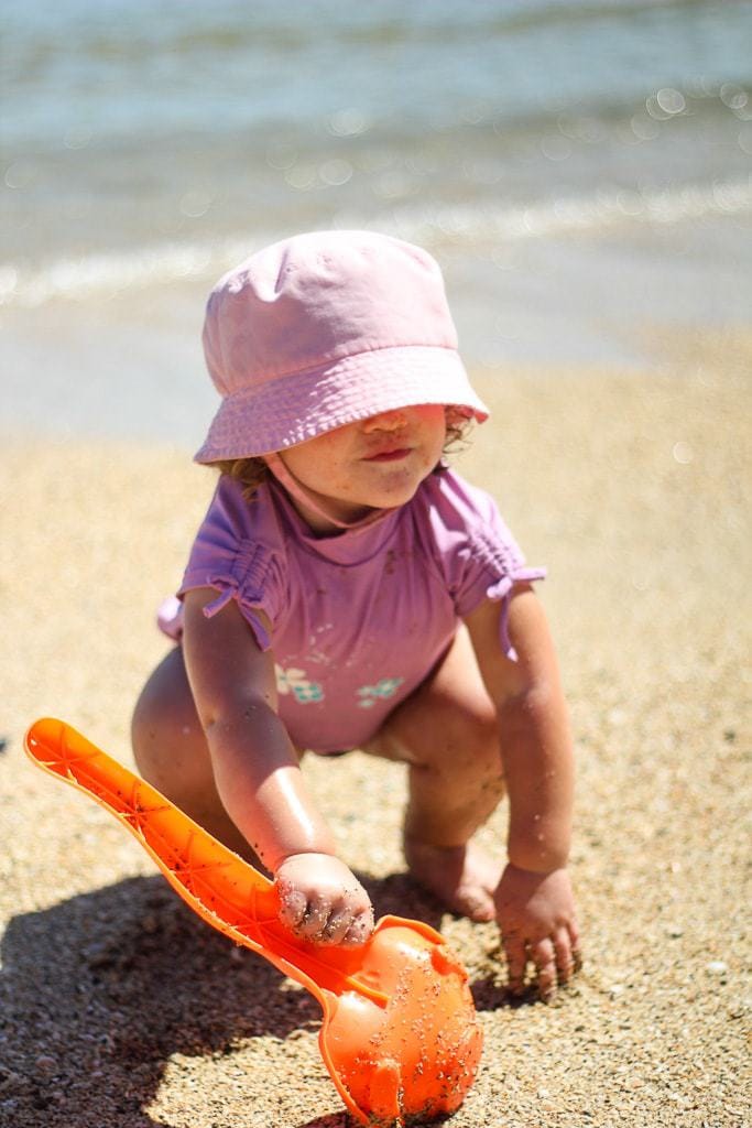Toddler girl wearing a pink outfit and digging in the sand with an orange scoop.