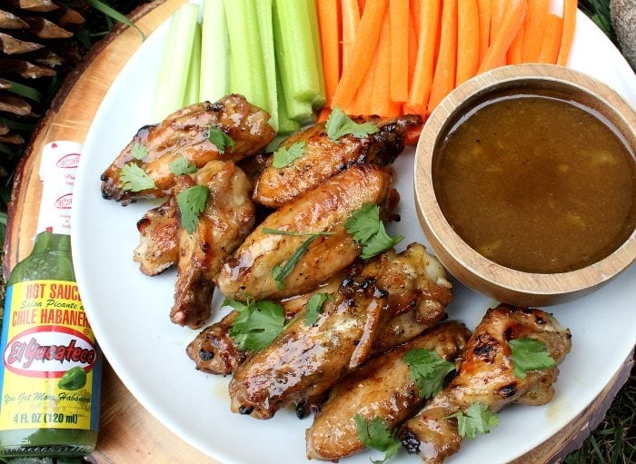Mango glazed chicken wings piled on a plate with a small bowl of dip and sliced vegetables.