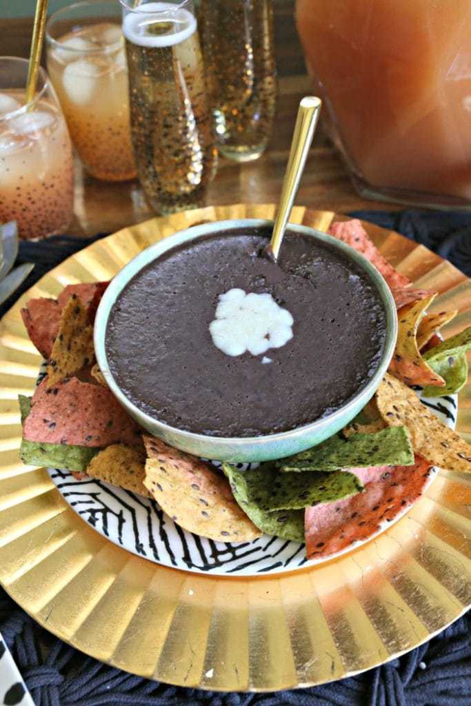 Black bean dip topped with cheese on a gold plate served with tri color tortilla chips.