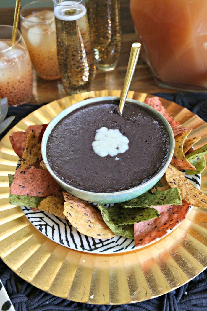 black bean dip topped with cheese on a gold plate served with tri color tortilla chips