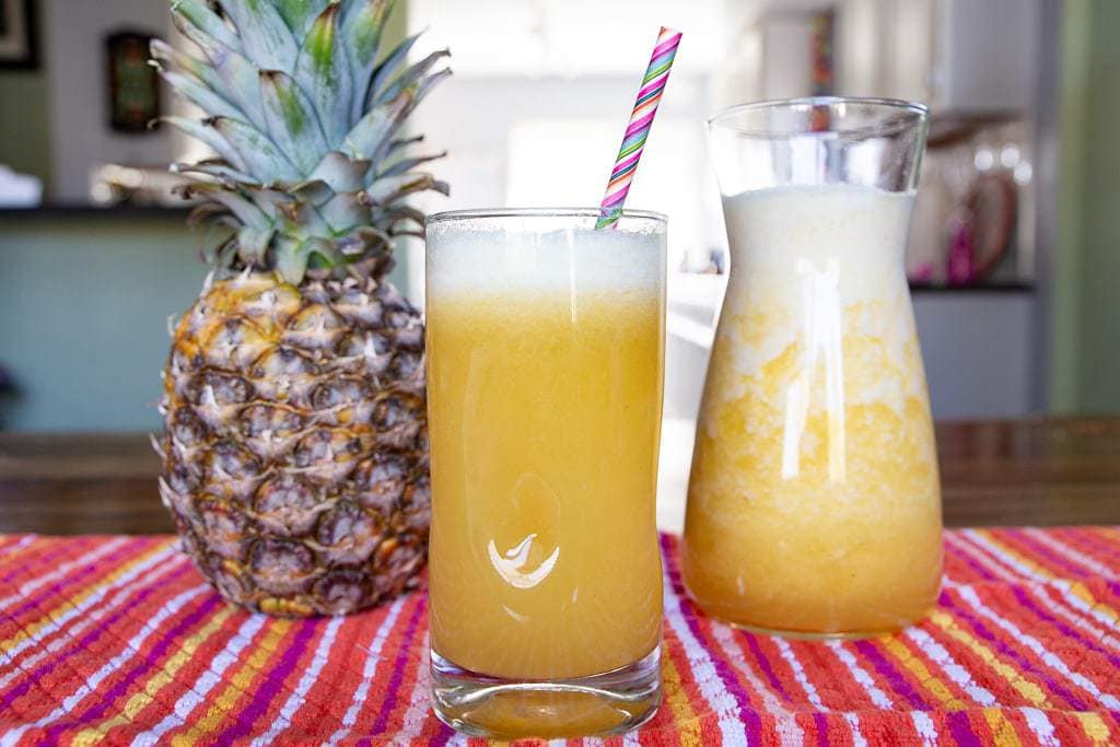 Pineapple fruit, glass of pineapple juice, and carafe placed on a striped cloth.