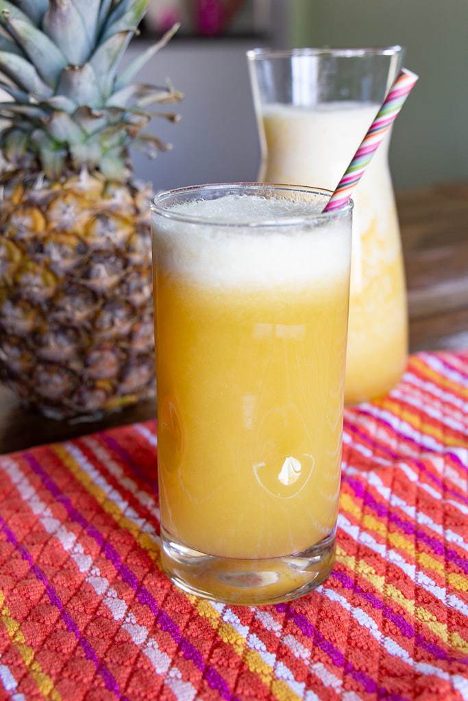 Fresh pineapple juice with a purple striped straw and a whole pineapple fruit in the background.
