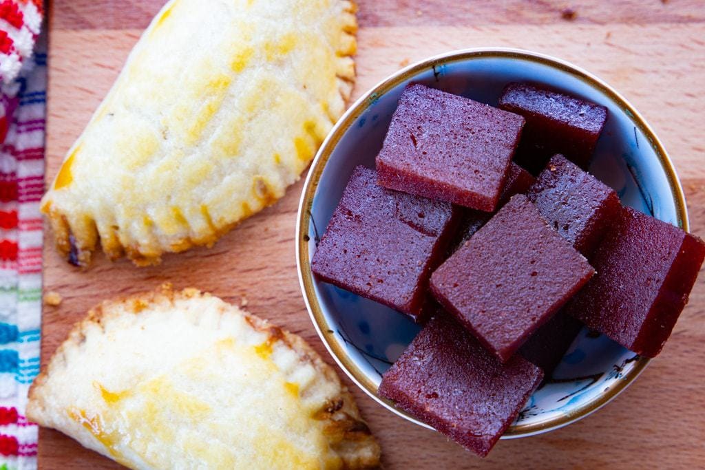 Two baked empanadas beside a bowl of guava paste sections.