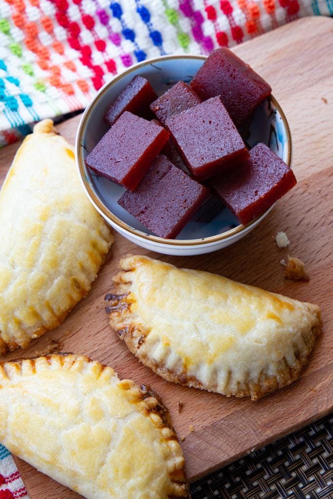 Small bowl containing chunks of guava paste next to three golden brown empanadas.
