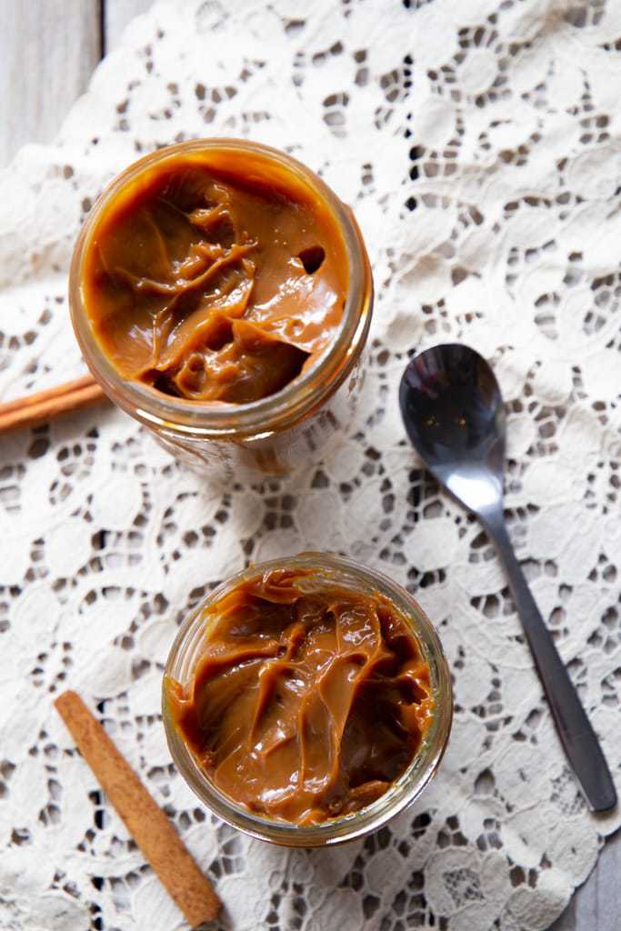 Aerial view of two jars of dulce de leche with cinnamon sticks and a spoon.